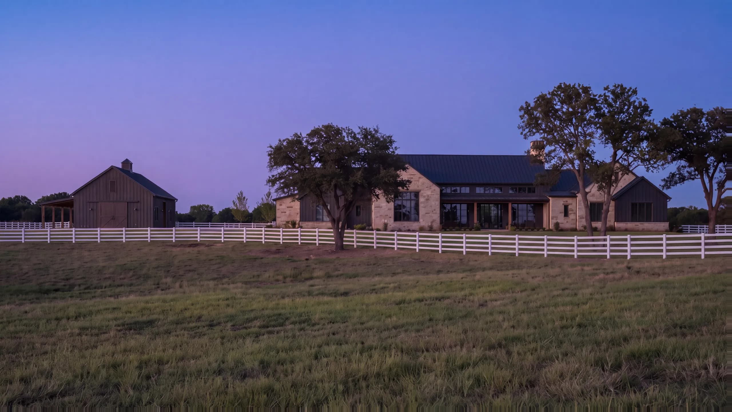 Limestone and dark board-and-batten farmhouse at dusk with lit windows, a barn beside it, scattered oaks and a white four-rail fence across the pasture