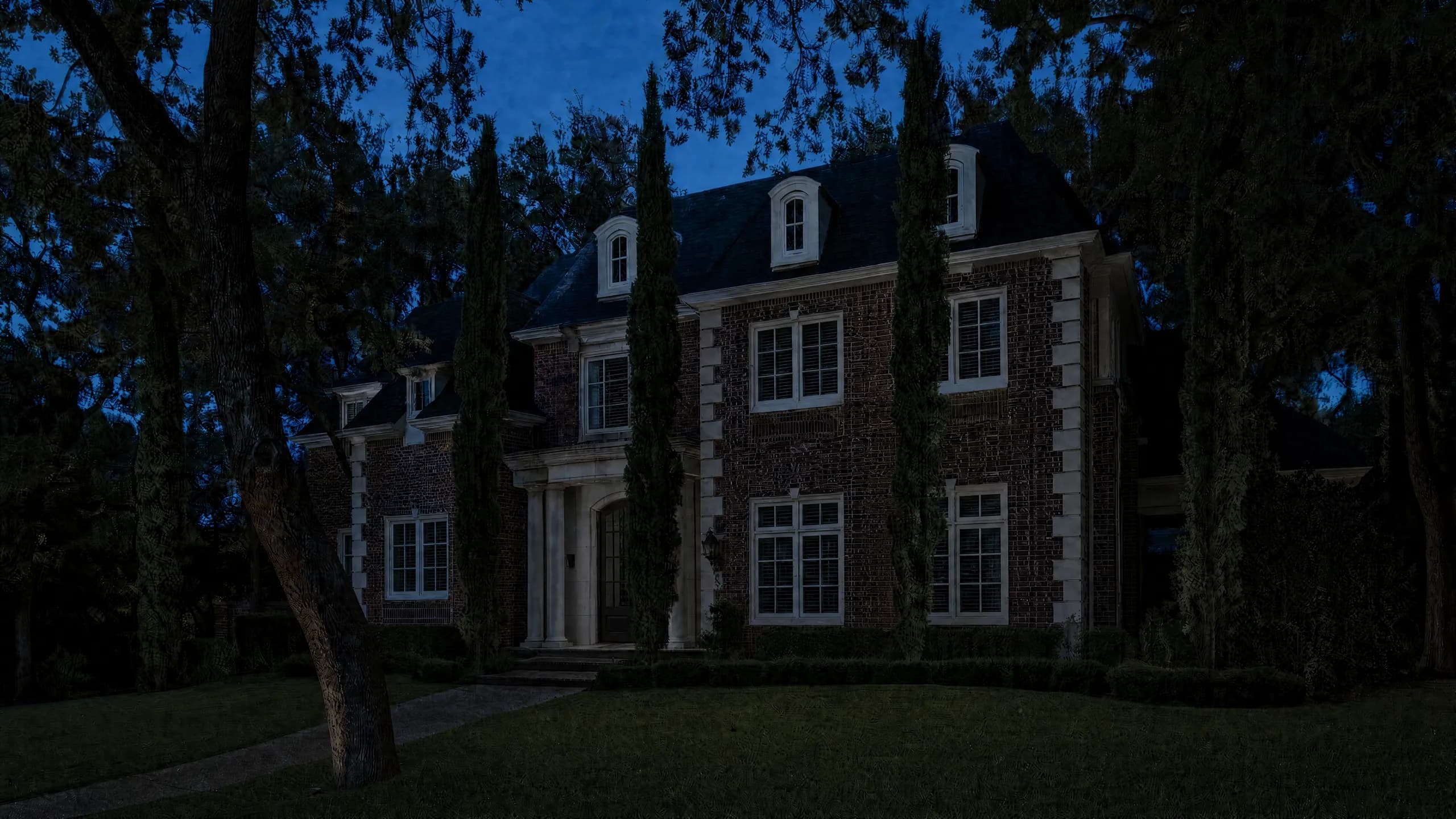 Red-brick home with arched dormers, limestone quoins and a columned entry at night, warm lit windows, uplit cypress and oaks, and a lit garden path