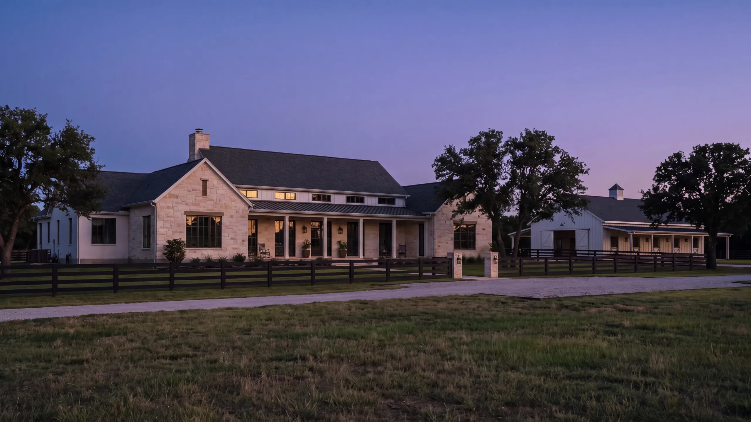 White-brick and limestone farmhouse at dusk behind a black four-rail fence, a gravel drive, a lit guest barn and spreading oaks