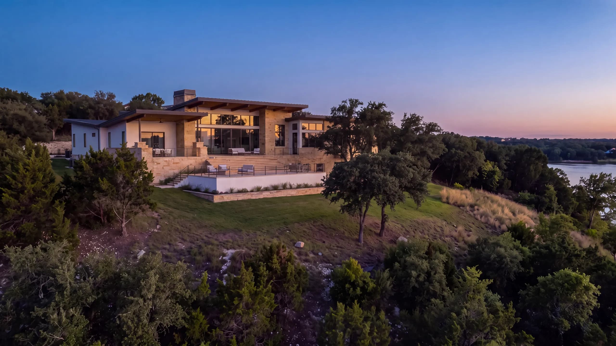 Modern stone and white-stucco home at dusk with a lit terrace on a hilltop above wooded slopes, a lake in the distance