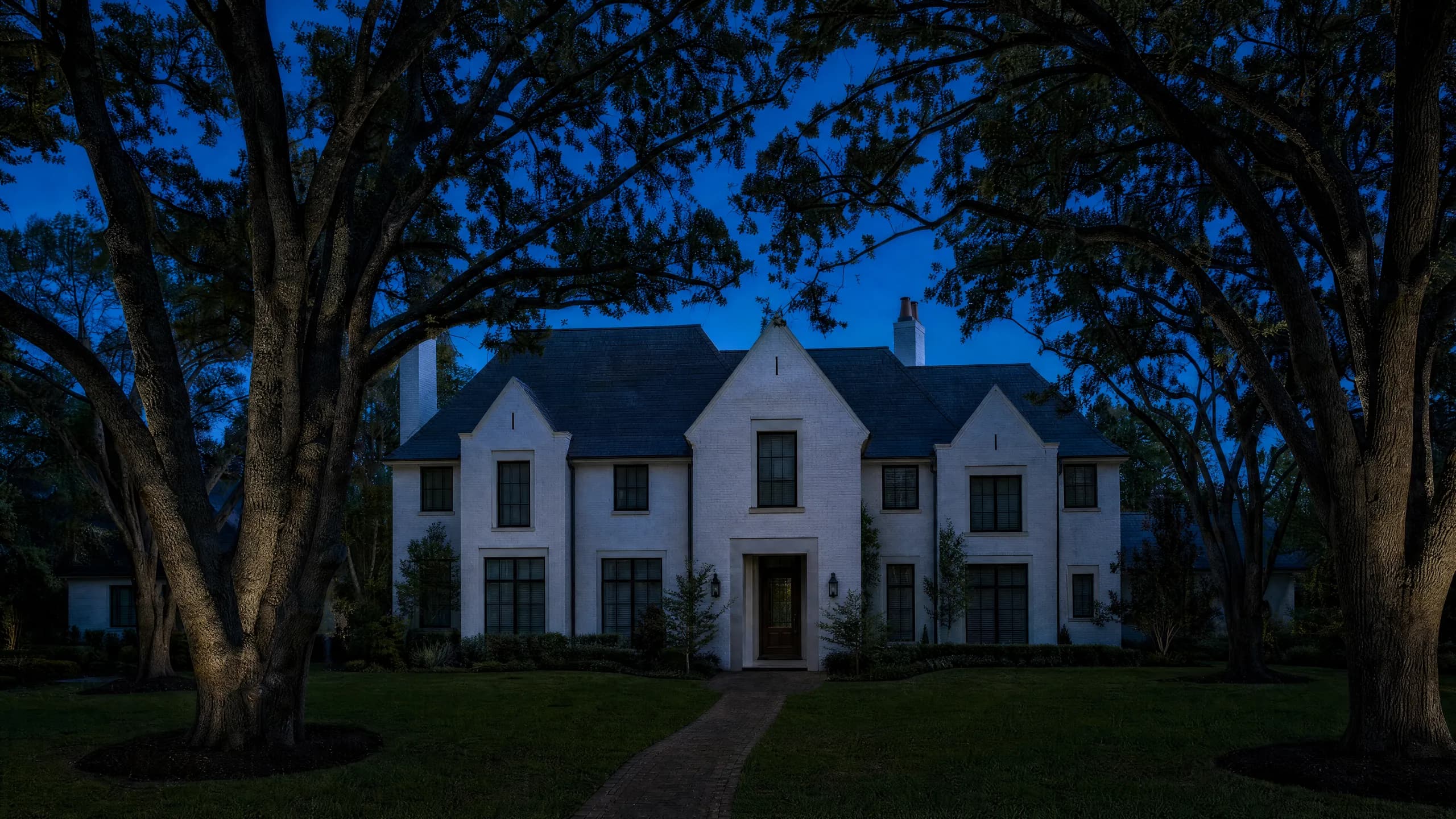 White-painted brick home at night beneath large uplit oaks, a brick walk lined with path lights leading to a lit entry