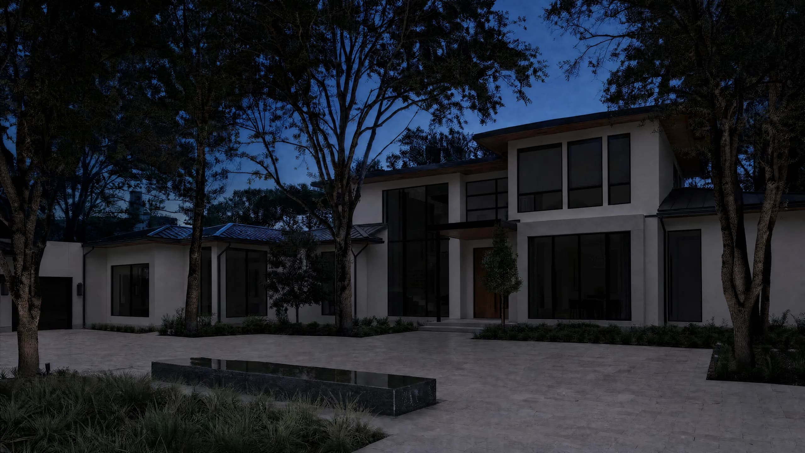 Modern white stucco and glass home at night with uplit trees, a still reflecting water feature and a stone-paved motor court