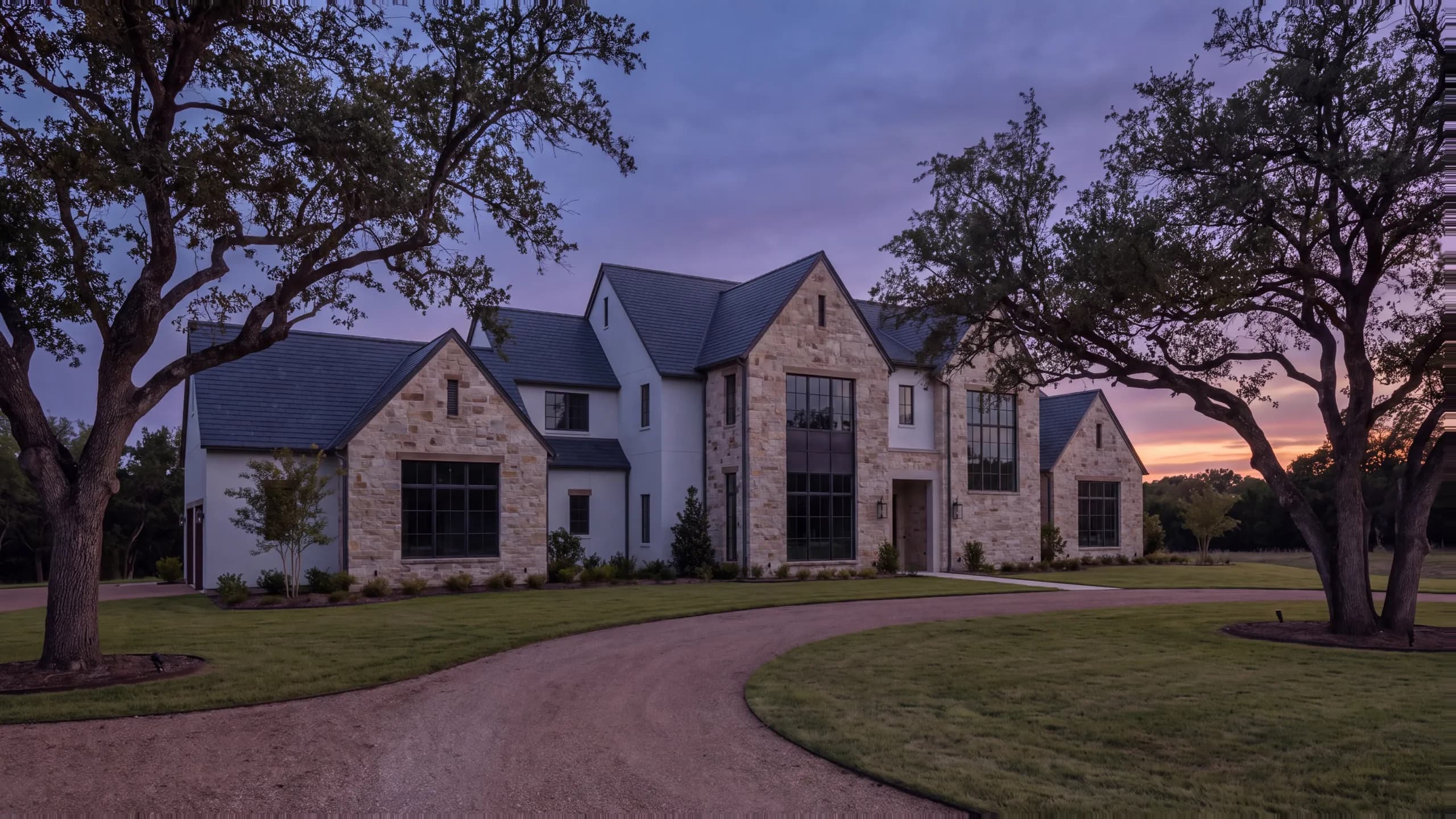Two-story limestone and white-brick manor at dusk with steep slate roofs, tall lit windows and an uplit facade, live oaks framing a curving gravel drive