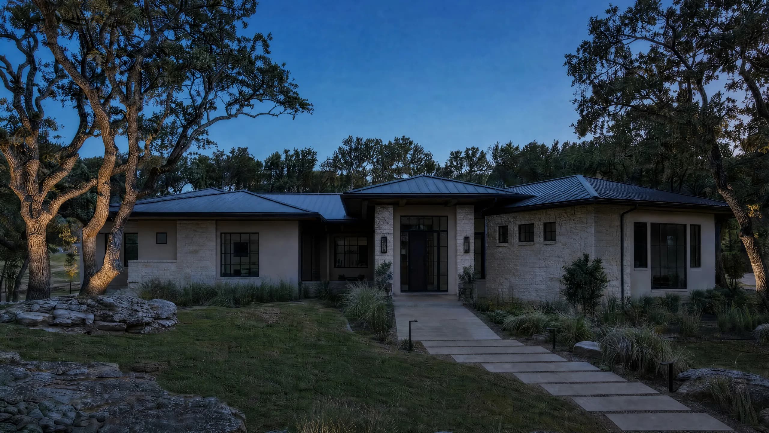 Low limestone and stucco home with a standing-seam metal roof at dusk, uplit oaks, path lights and a stepping-stone walk to a lit entry