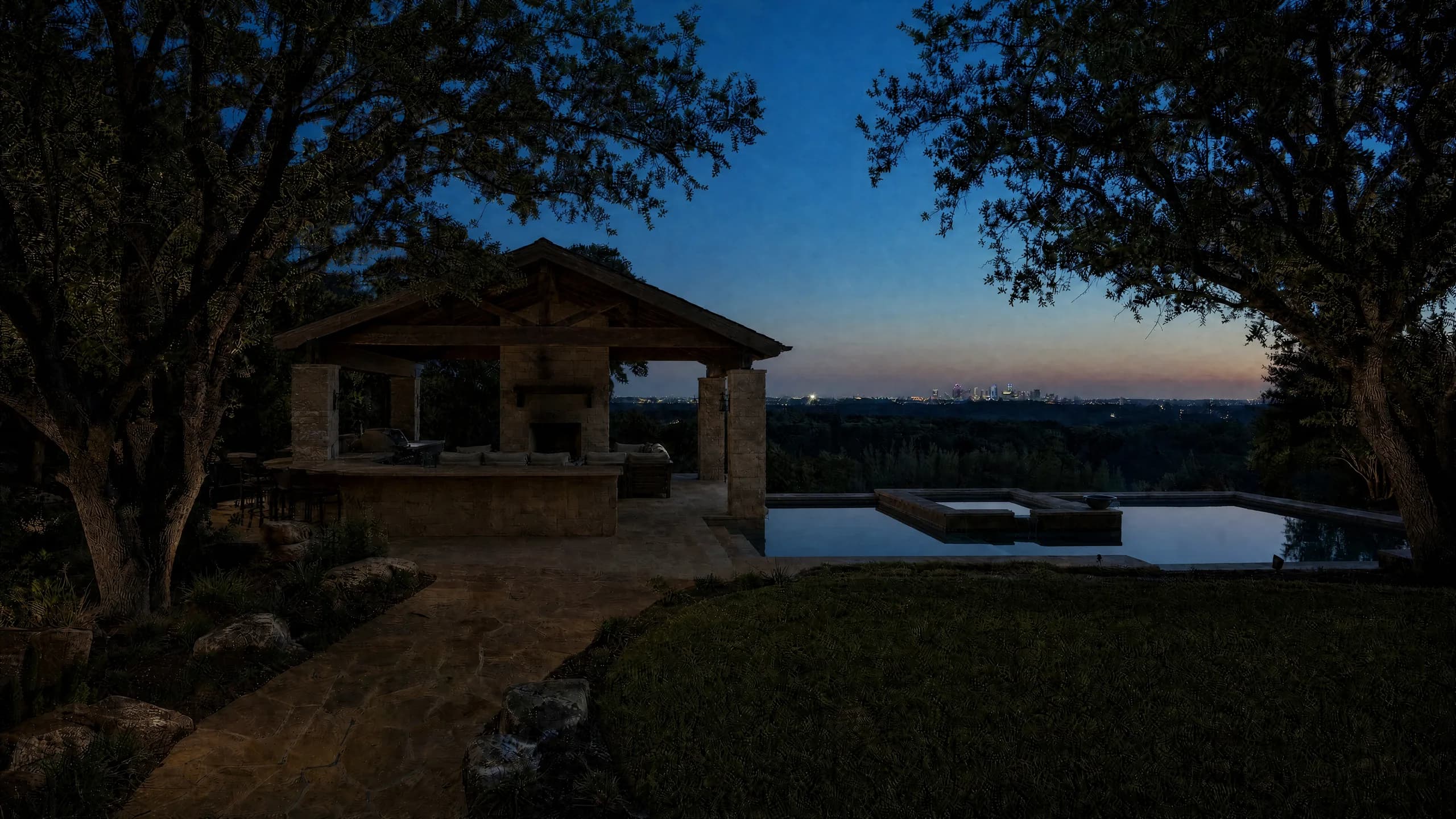 Stone outdoor pavilion at dusk beside a lit pool, with uplit oaks and a distant skyline