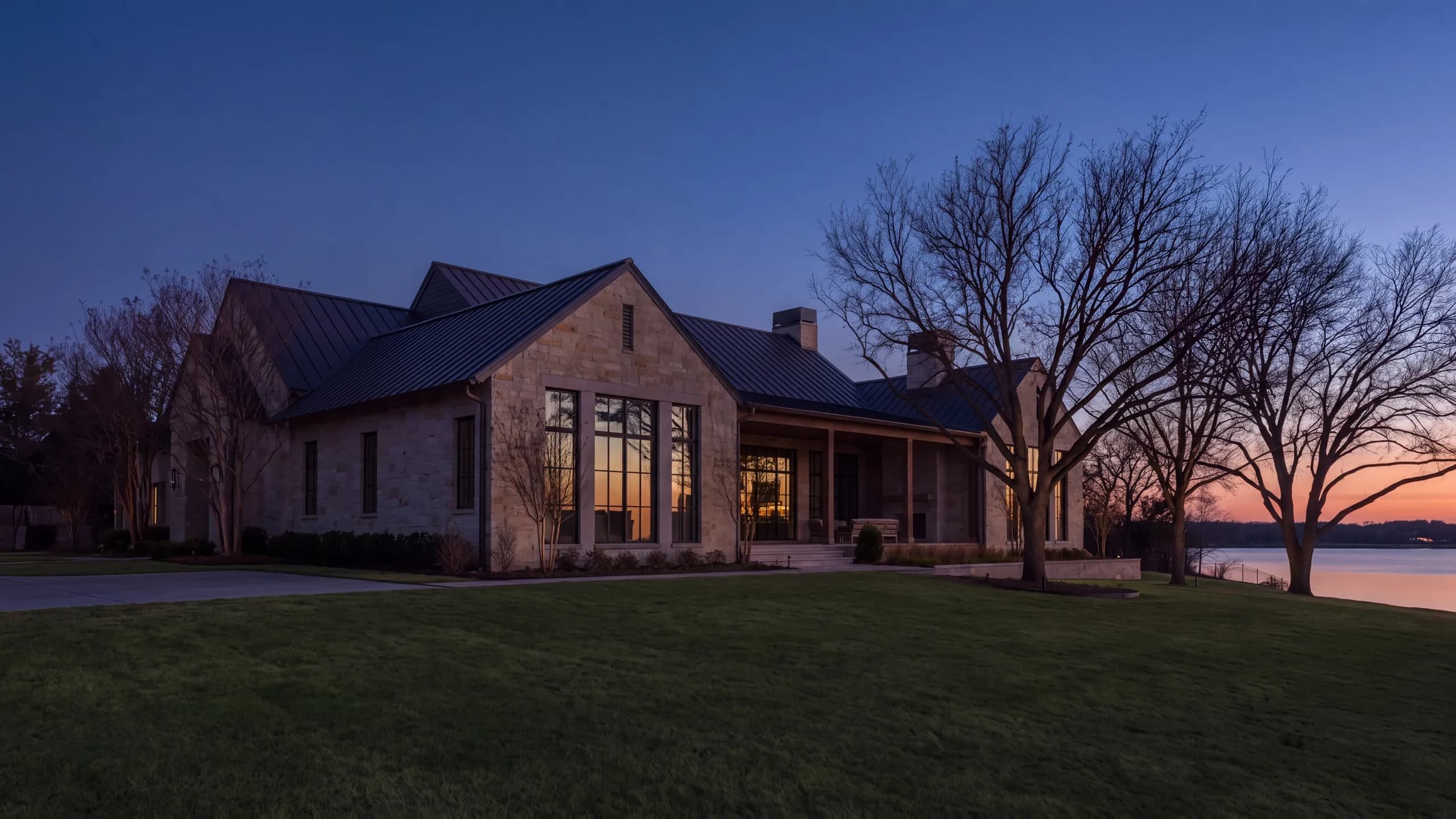 Limestone farmhouse at dusk under a standing-seam roof, tall lit windows and a deep porch, bare oaks across the lawn and water beyond