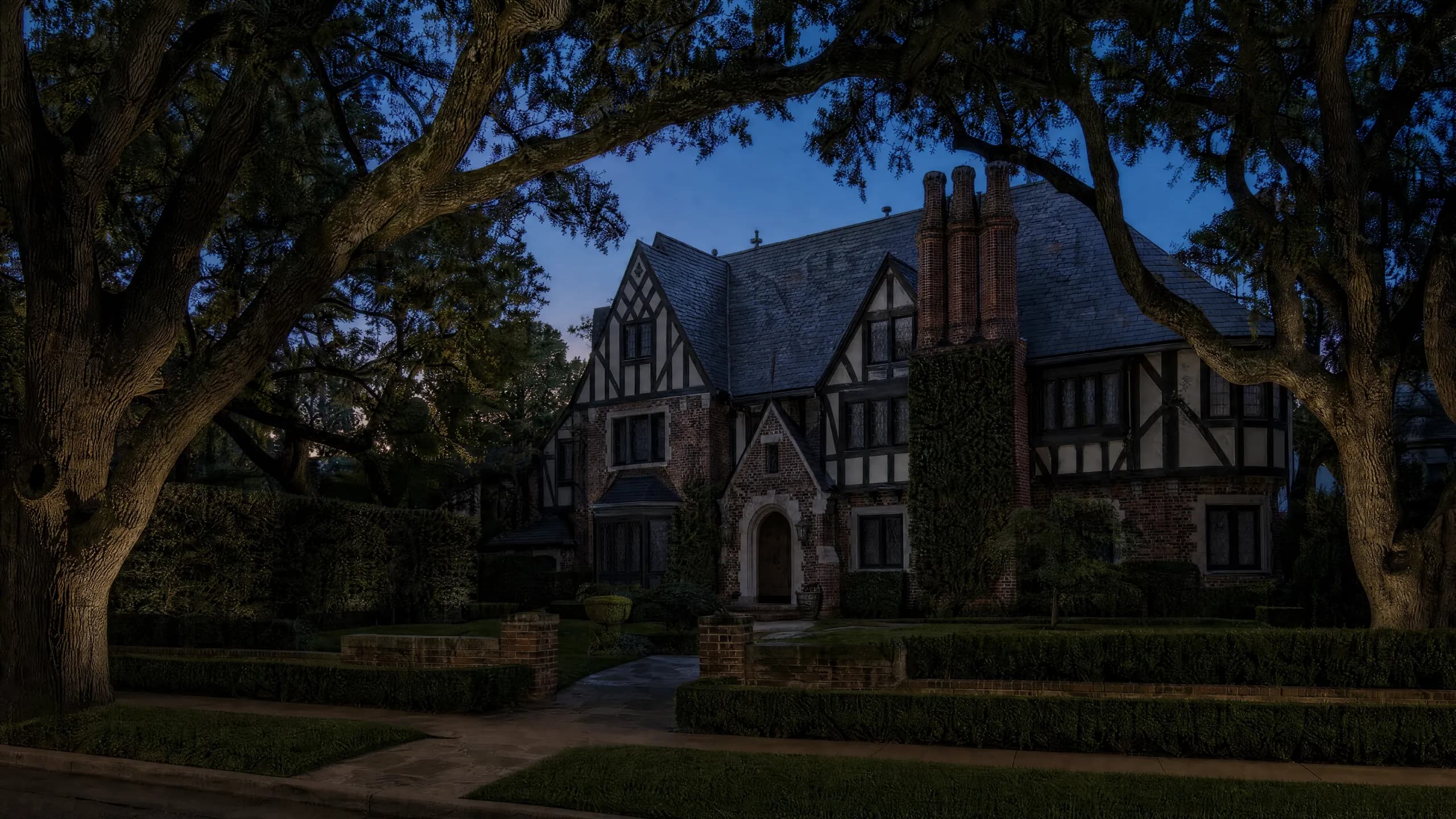 Tudor Revival home at dusk with half-timbered gables, uplit oaks and a hedge-lined walk