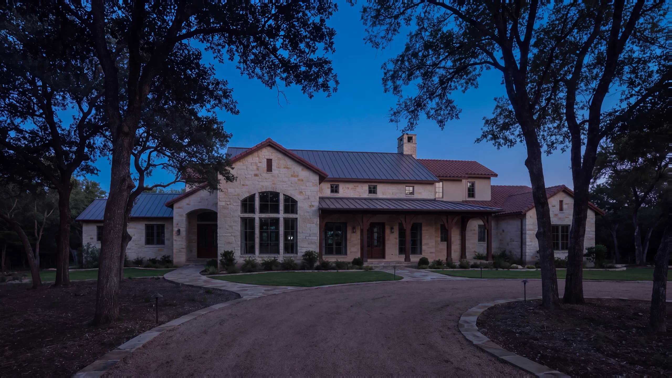 Single-story limestone ranch home at dusk with a standing-seam metal roof, a long timber porch and an arched window, path lights along a curving drive beneath large oaks