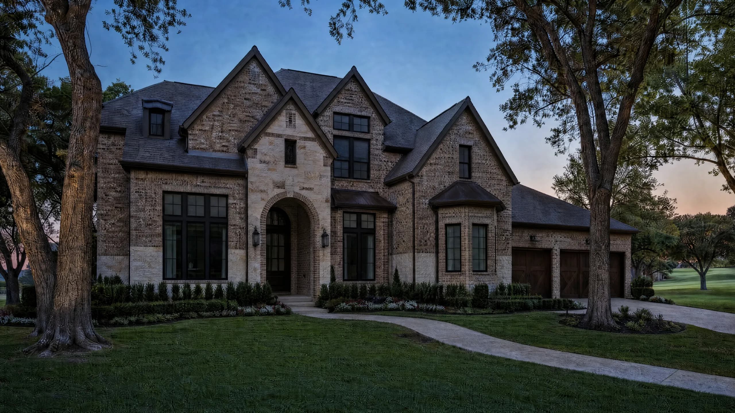 Brick and limestone home with steep gables and an arched entry at dusk, lantern sconces, path lights and uplit trees over a manicured lawn