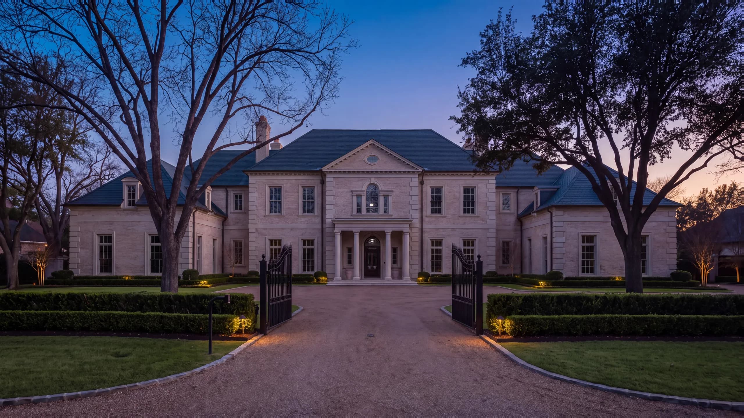Limestone Georgian manor at dusk with a columned portico, lit windows and black iron gates open onto a gravel motor court beneath tall oaks