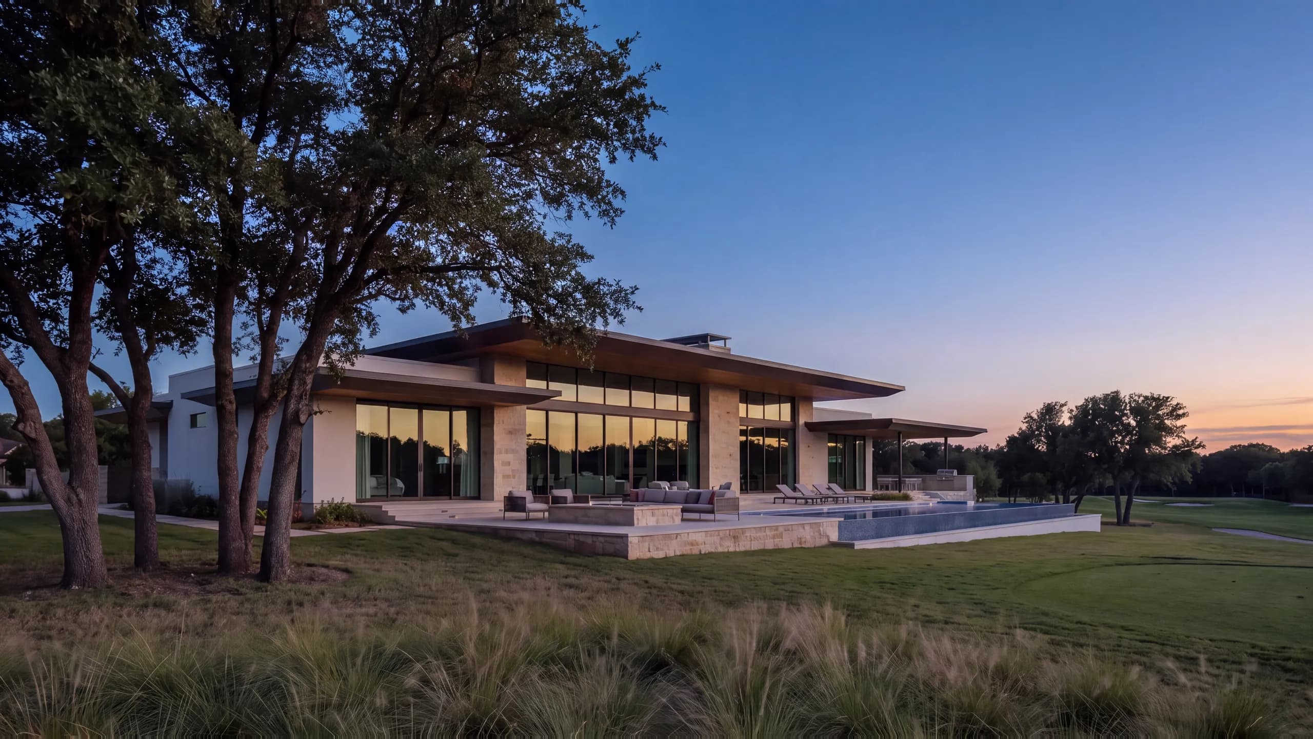 Low modern stone and glass home at dusk under a deep cantilevered roof, a lit great room, a fire table on the terrace and an edge pool, with a fairway and oaks beyond