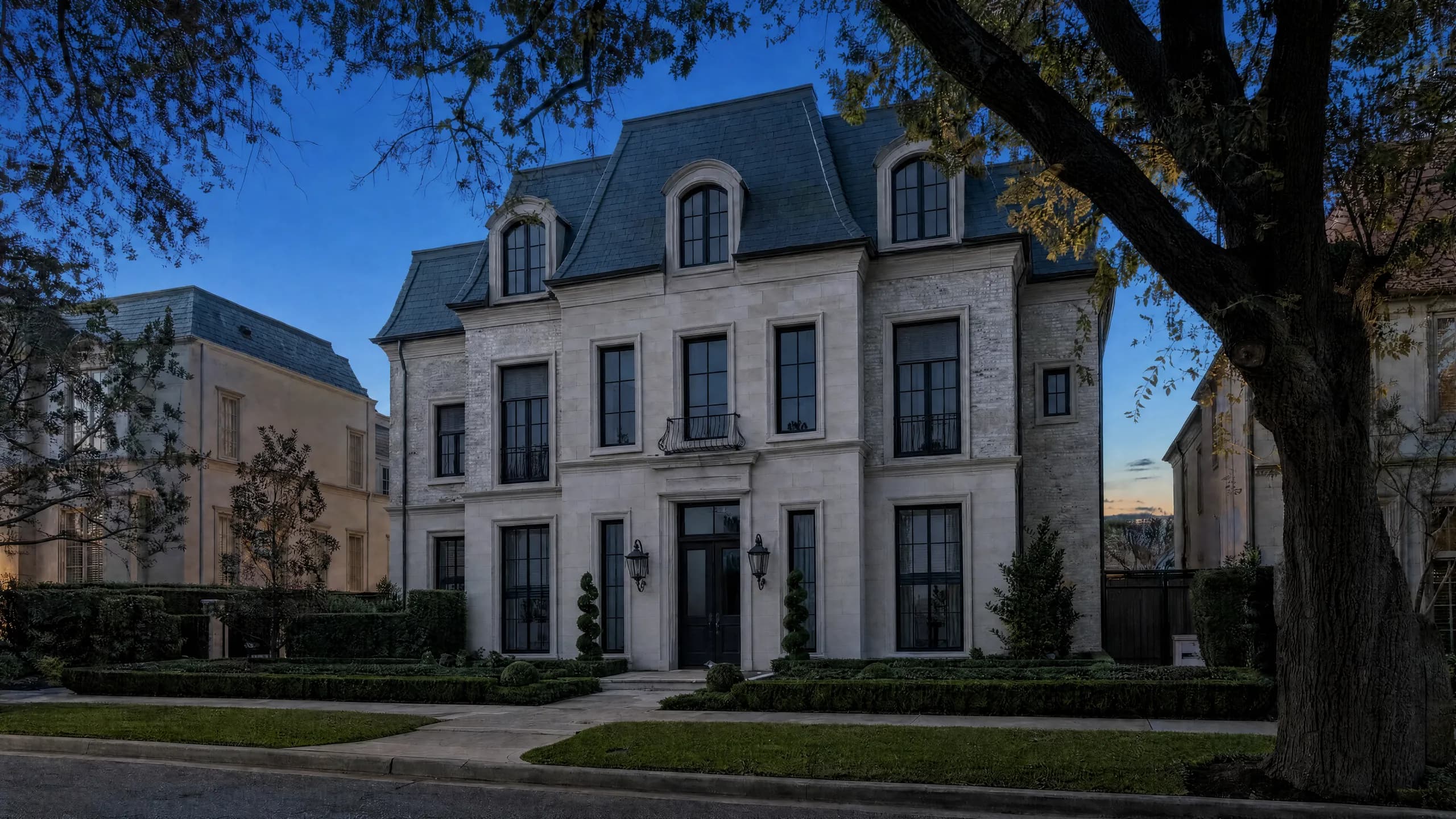 Three-story French limestone home at dusk on a narrow lot with lanterns and boxwood parterre