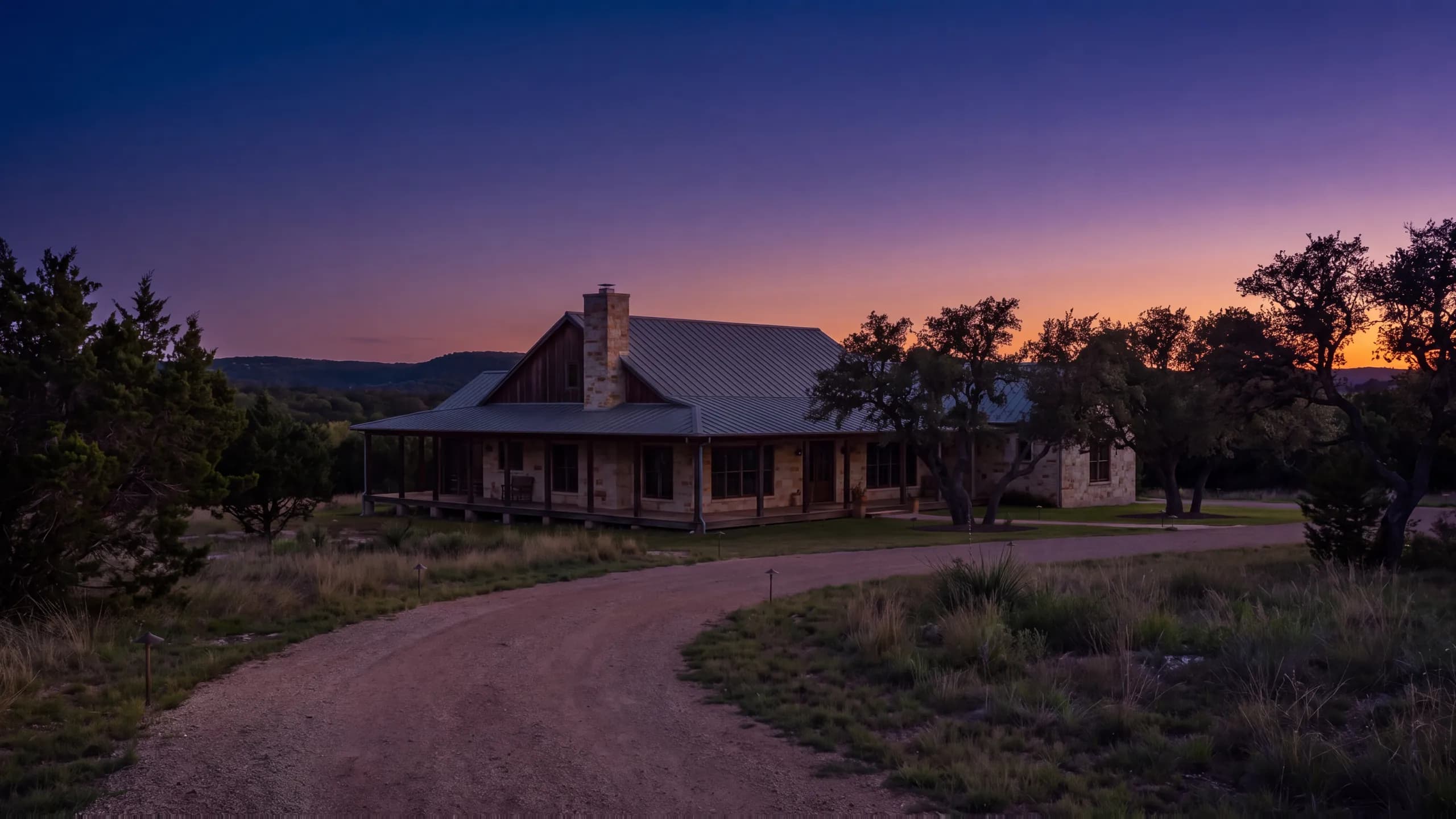 Stone ranch house with a deep porch and metal roof at dusk, path lights along a curving gravel drive through native grass, oaks and low hills beyond