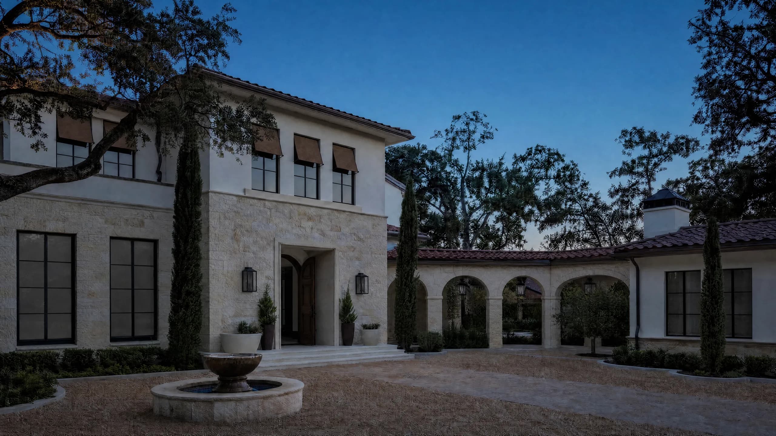 Limestone and stucco courtyard home at dusk with lantern sconces, an arcaded walk, a stone fountain and uplit cypress and oaks