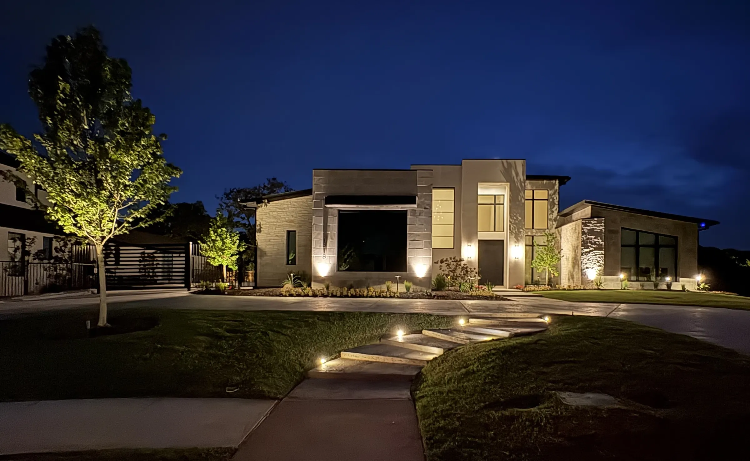 Contemporary stone residence in Southlake at dusk, with uplit trees and facade, and a lit stepping-stone walk across the lawn