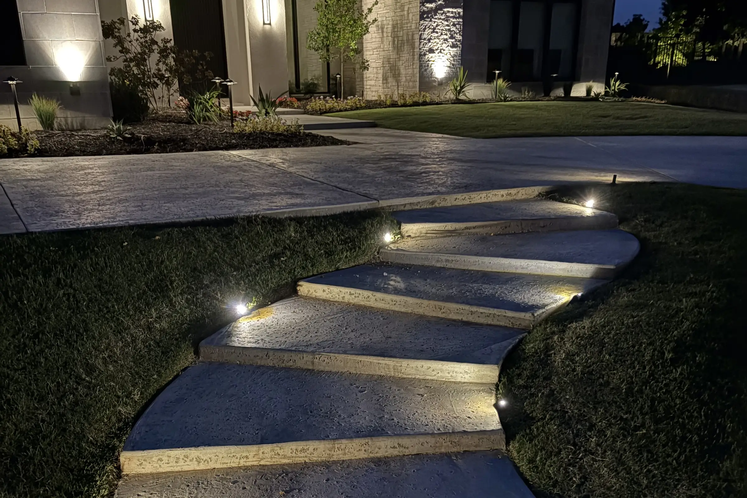 Low lights along stone steps curving up a lawn at dusk to a front walk and planted beds
