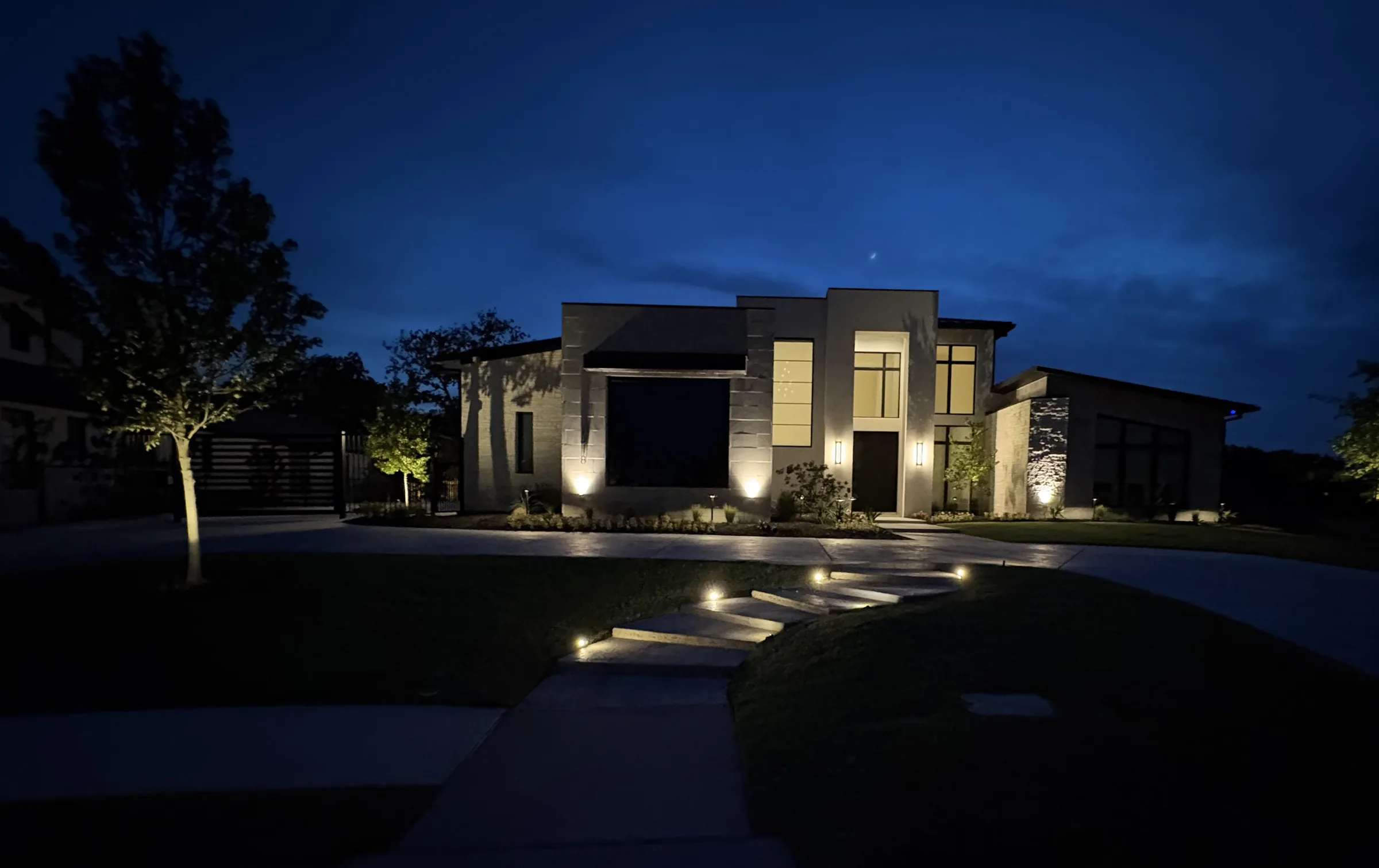 Modern stone residence at dusk, with uplit facade and trees and lit steps rising across the lawn to the drive