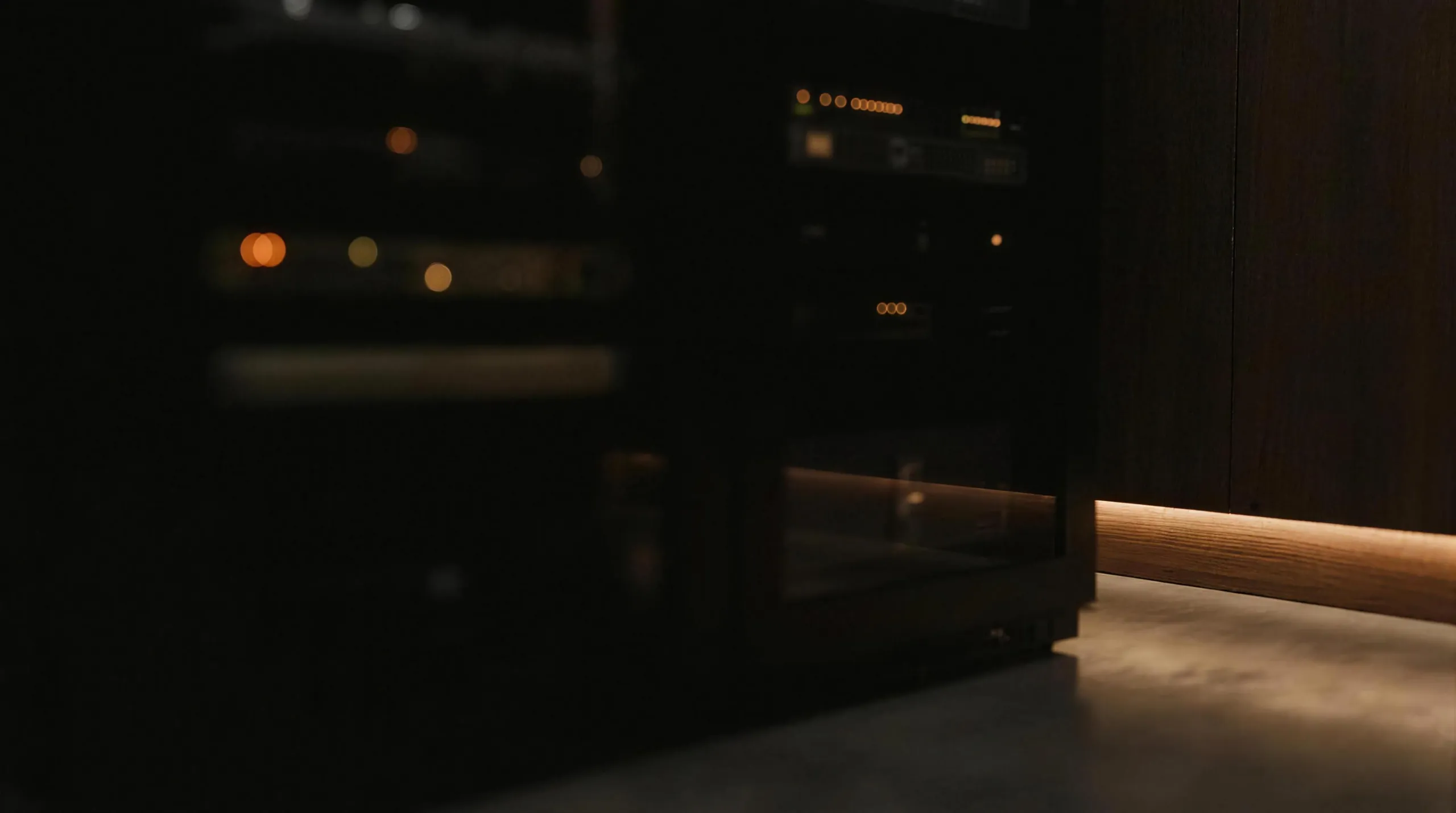 An equipment room in deep shadow — soft glow of indicator lights and warm cove lighting on walnut
