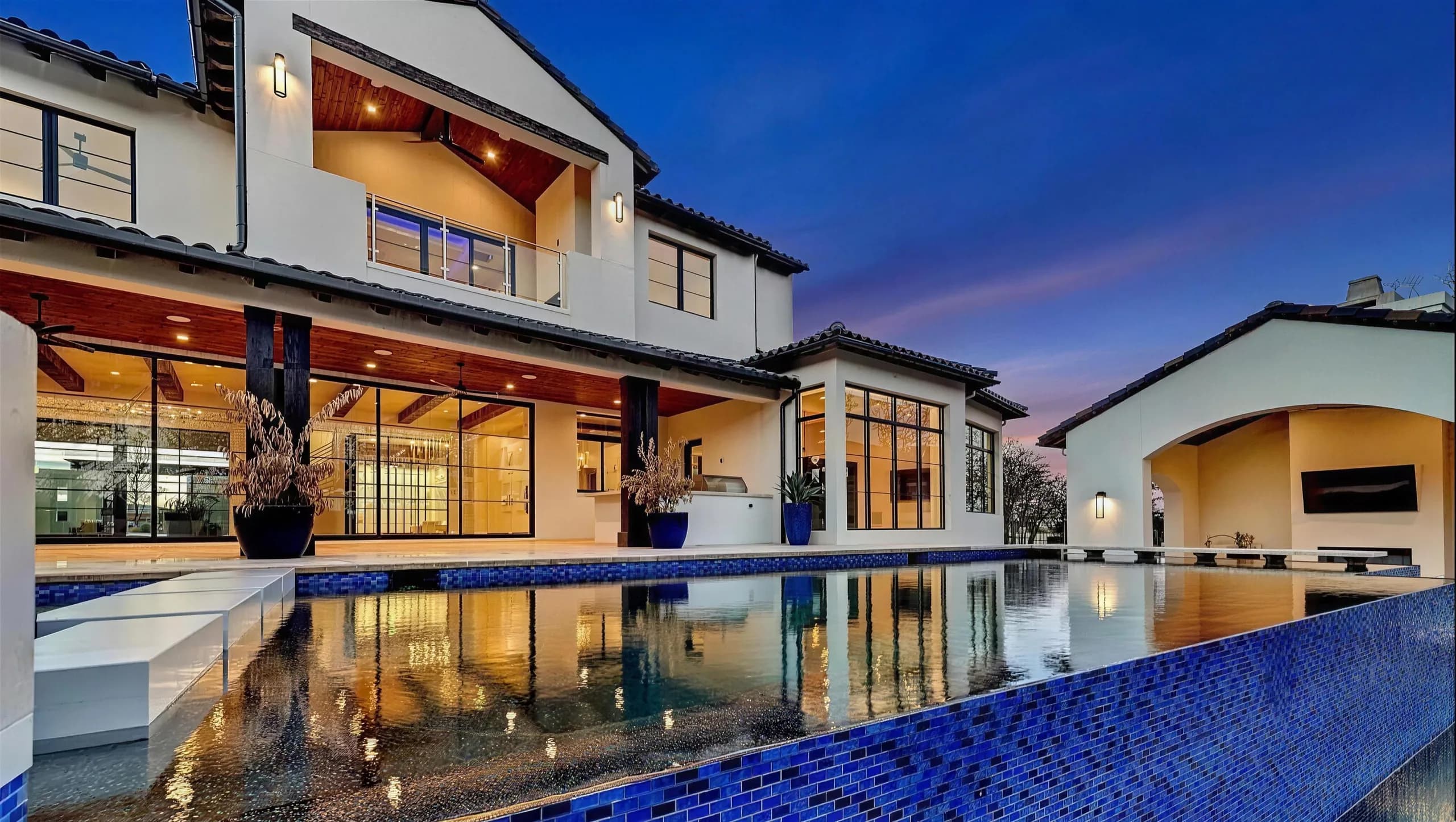 The back of the home reflected in a blue-tiled zero-edge pool at dusk, the upstairs balcony lit beneath a cedar ceiling and the cabana at the far end