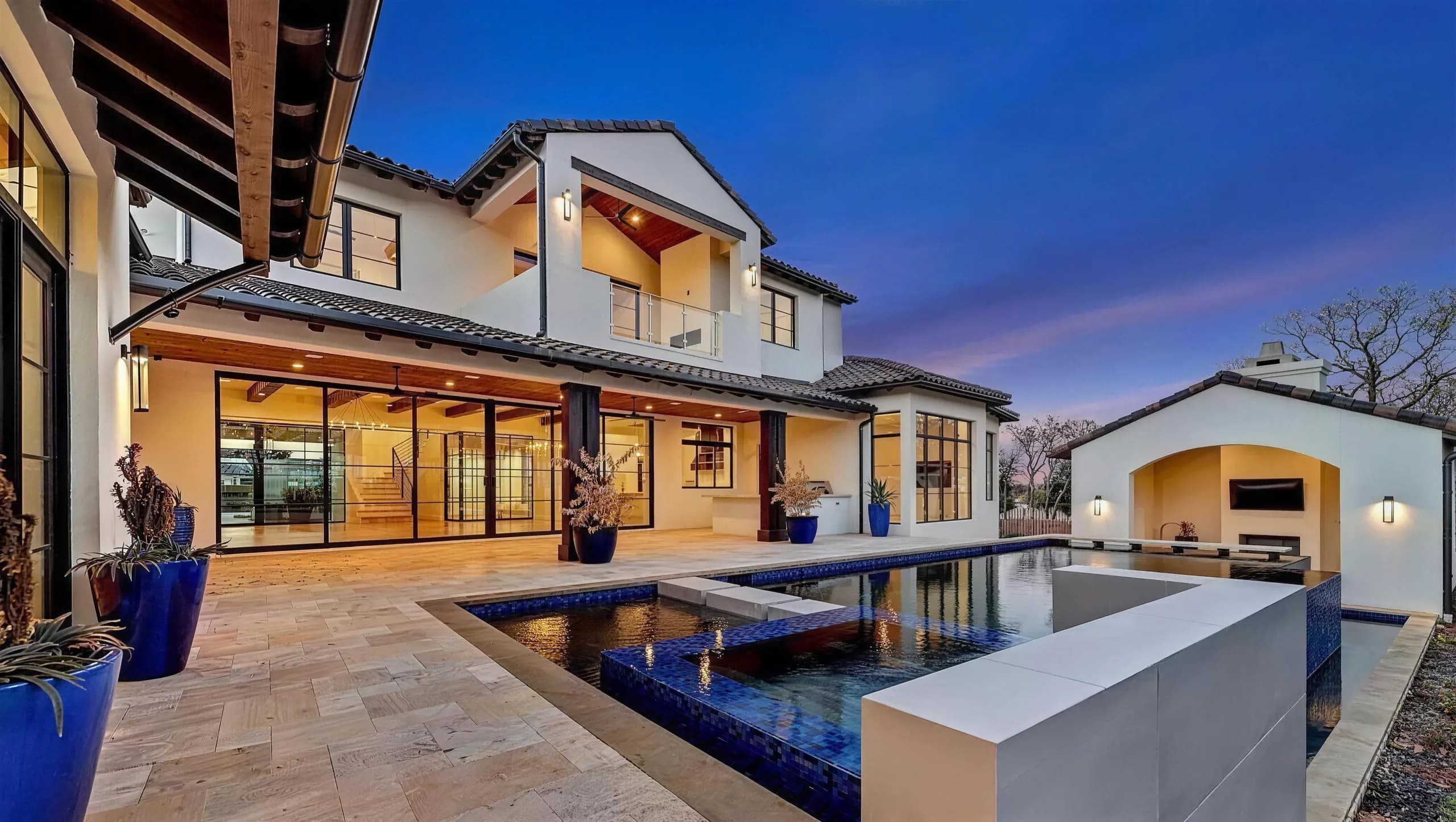 Two-story white stucco home with a tile roof at dusk, its glass walls lit warm across a stone patio, with a blue-tiled pool and an arched cabana beside it